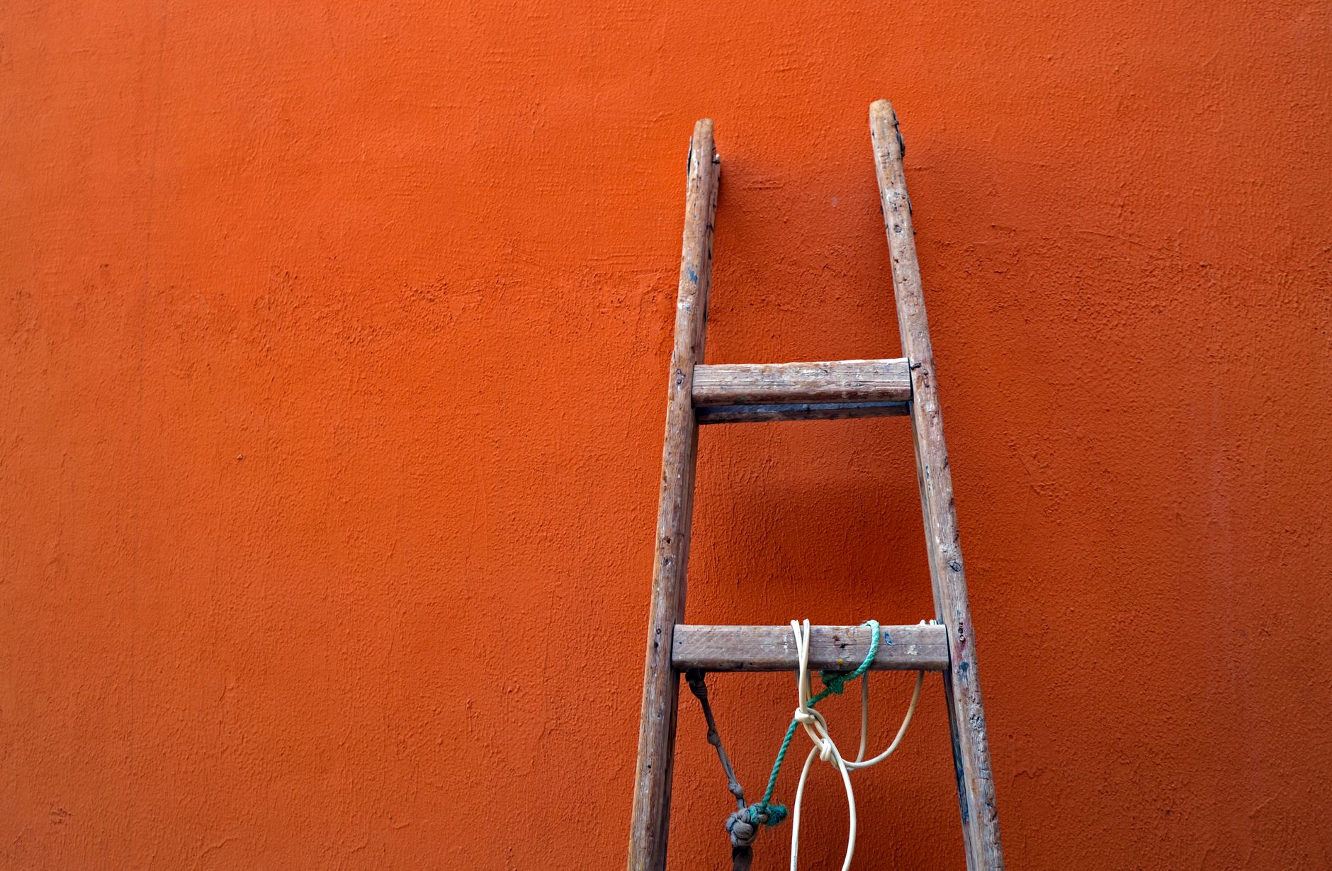 A wooden ladder leaning against an orange wall, tied with rope.
